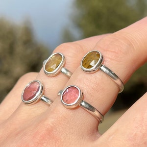 May include: Four silver rings with oval-shaped gemstones. Two rings feature a light pink gemstone, and two rings feature a yellow-brown gemstone.