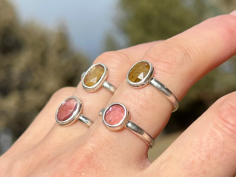 May include: Four silver rings with oval-shaped gemstones. Two rings feature a light pink gemstone, and two rings feature a yellow-brown gemstone.