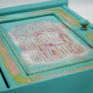 May include: A turquoise wooden box with a framed front featuring the text "HOME SWEET HOME" in a decorative font. The frame has a rope-like border and is accented with pink and green paint. A small, round knob is on the side.