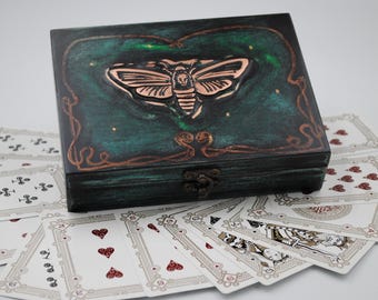 May include: A dark green wooden box with a copper-colored moth design on the lid, resting on a fan of playing cards. The cards have red hearts and ornate designs. The box has a clasp closure.