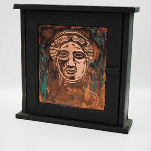 May include: A small, black wooden cabinet featuring a copper relief of a classical female face. The cabinet is rectangular with a door and a dark, weathered finish. The copper relief depicts a woman's face in intricate detail, with a classical theme.
