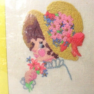 May include: Embroidered picture of a girl with brown hair wearing a yellow straw hat with pink and blue flowers and a red bow. She is holding a bouquet of pink and blue flowers.