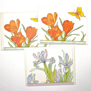 May include: Three greeting cards with floral designs. The first card features orange crocuses with green leaves and a yellow butterfly. The second card features purple irises with green leaves. The third card features orange crocuses with green leaves and a yellow butterfly.