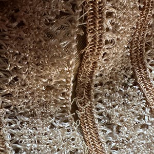 May include: Close-up of a textured, woven fabric in shades of beige and tan. The design features a combination of looped and braided elements, creating a tactile and intricate pattern. The overall effect is a soft, neutral color palette.