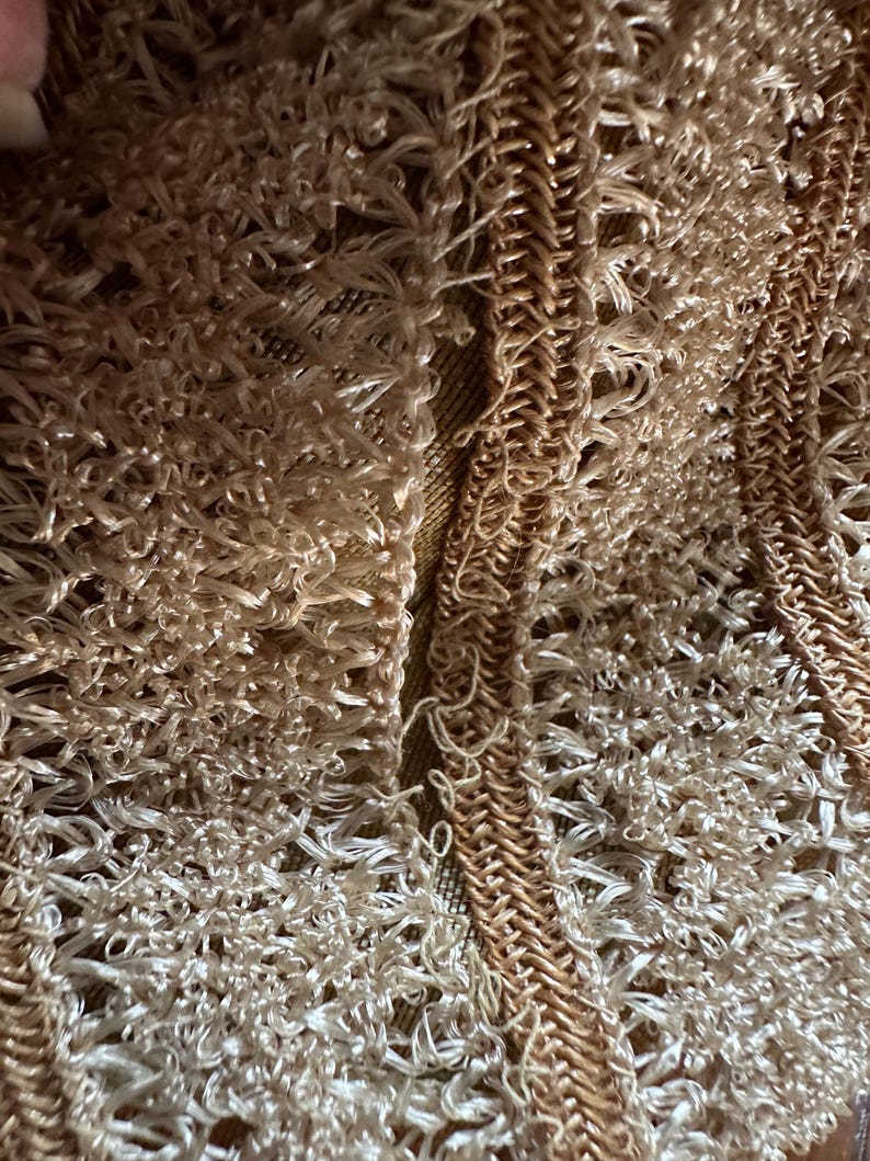 May include: Close-up of a textured, woven fabric in shades of beige and tan. The design features a combination of looped and braided elements, creating a tactile and intricate pattern. The overall effect is a soft, neutral color palette.
