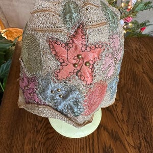 May include: A vintage-style hat with a star-shaped patch and various fabric patches in pink, blue, and green. The hat is embellished with gold-colored beads and a gold-colored trim. The hat is displayed on a light yellow stand.