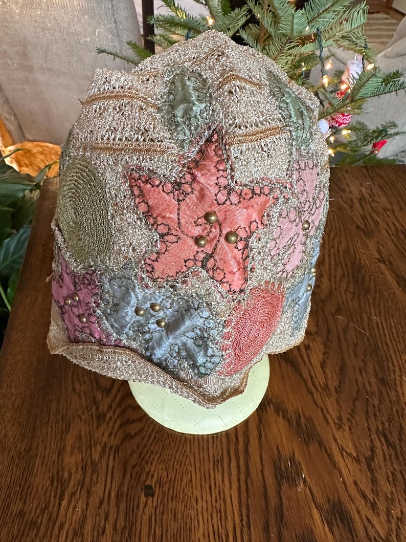 May include: A vintage-style hat with a star-shaped patch and various fabric patches in pink, blue, and green. The hat is embellished with gold-colored beads and a gold-colored trim. The hat is displayed on a light yellow stand.