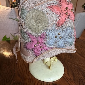 May include: An antique hat with a patchwork design. The hat features a mix of pastel colors, including pink, blue, and green, with gold accents and embellishments. It is displayed on a cream-colored stand with a yellow bow.