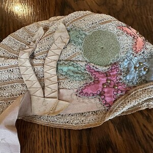 May include: An antique beige lace bonnet with a patchwork design. The bonnet features a star and circular design in pink, green, and blue, adorned with small gold beads. Beige ribbons are attached to the bonnet.