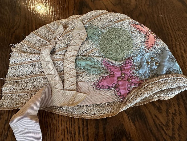 May include: An antique beige lace bonnet with a patchwork design. The bonnet features a star and circular design in pink, green, and blue, adorned with small gold beads. Beige ribbons are attached to the bonnet.