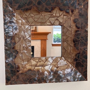 May include: A square wall mirror featuring a wide, decorative frame composed of numerous small, triangular metal pieces. The frame displays a gradient of dark, scorched brown and metallic bronze tones with a textured, uneven surface finish.