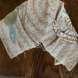 May include: A vintage, beige lace collar with intricate openwork and decorative trim. The collar features a circular embellishment and accents of pale green and pink ribbon. The delicate fabric is displayed on a wooden surface.