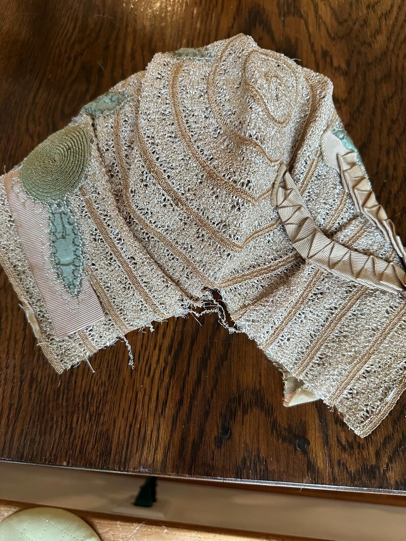May include: A vintage, beige lace collar with intricate openwork and decorative trim. The collar features a circular embellishment and accents of pale green and pink ribbon. The delicate fabric is displayed on a wooden surface.