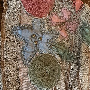 May include: Close-up of a vintage textile with intricate details. Features a beige crocheted background with appliqued shapes in pink, blue, and green. Gold beads and a spiral design add texture and visual interest. A unique, handcrafted piece.