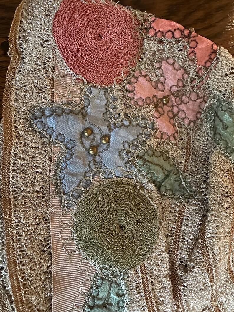 May include: Close-up of a vintage textile with intricate details. Features a beige crocheted background with appliqued shapes in pink, blue, and green. Gold beads and a spiral design add texture and visual interest. A unique, handcrafted piece.