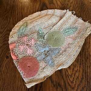 May include: An antique, beige-colored lace cap with floral embellishments. The cap features appliqued flowers in pink, blue, and green, with gold-colored beads. The edges are trimmed with delicate lace, and the cap rests on a wooden surface.