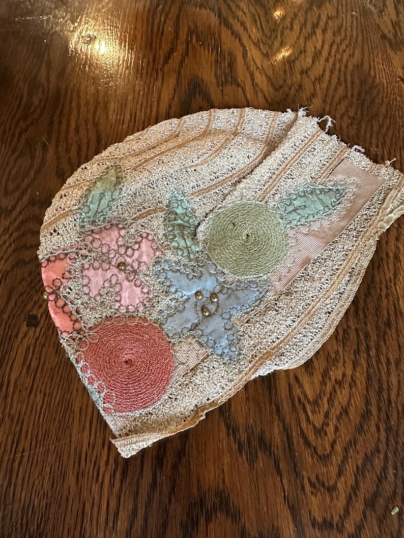 May include: An antique, beige-colored lace cap with floral embellishments. The cap features appliqued flowers in pink, blue, and green, with gold-colored beads. The edges are trimmed with delicate lace, and the cap rests on a wooden surface.