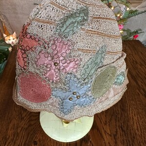 May include: A vintage cloche hat with a gold-colored crocheted crown and colorful fabric embellishments. The hat features pink, blue, and green floral designs, accented with small gold beads. The brim is trimmed with a gold-colored scalloped edge.