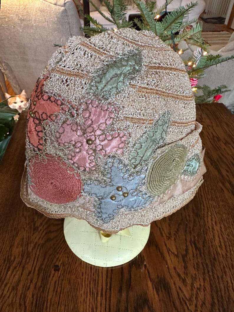 May include: A vintage cloche hat with a gold-colored crocheted crown and colorful fabric embellishments. The hat features pink, blue, and green floral designs, accented with small gold beads. The brim is trimmed with a gold-colored scalloped edge.