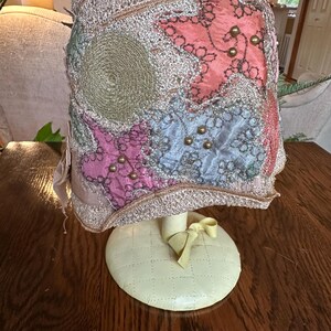 May include: A vintage hat with a decorative design. The hat features a mix of colors, including pink, blue, and gold, with star-shaped and circular embellishments. It is displayed on a cream-colored stand with a bow.