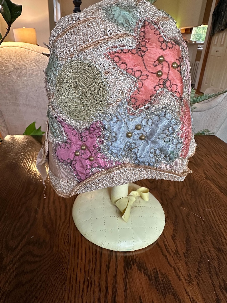 May include: A vintage hat with a decorative design. The hat features a mix of colors, including pink, blue, and gold, with star-shaped and circular embellishments. It is displayed on a cream-colored stand with a bow.