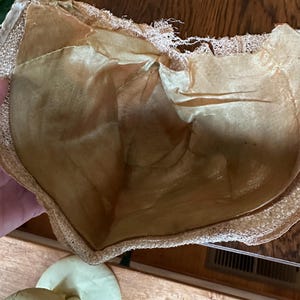 May include: Vintage beige bra with a delicate lace trim and a satin lining. The bra appears to be made of a soft, possibly silk, material. The image also shows a pair of matching quilted pads.