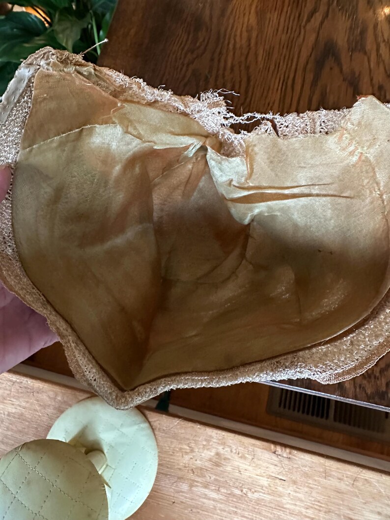 May include: Vintage beige bra with a delicate lace trim and a satin lining. The bra appears to be made of a soft, possibly silk, material. The image also shows a pair of matching quilted pads.