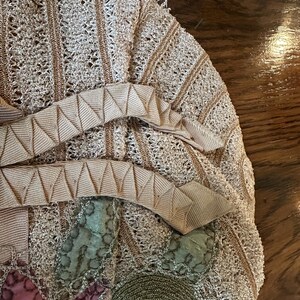 May include: Close-up of a vintage beige crocheted handbag with a decorative ribbon and floral accents. The bag features a detailed openwork design with a light brown trim and a pink and green floral pattern.