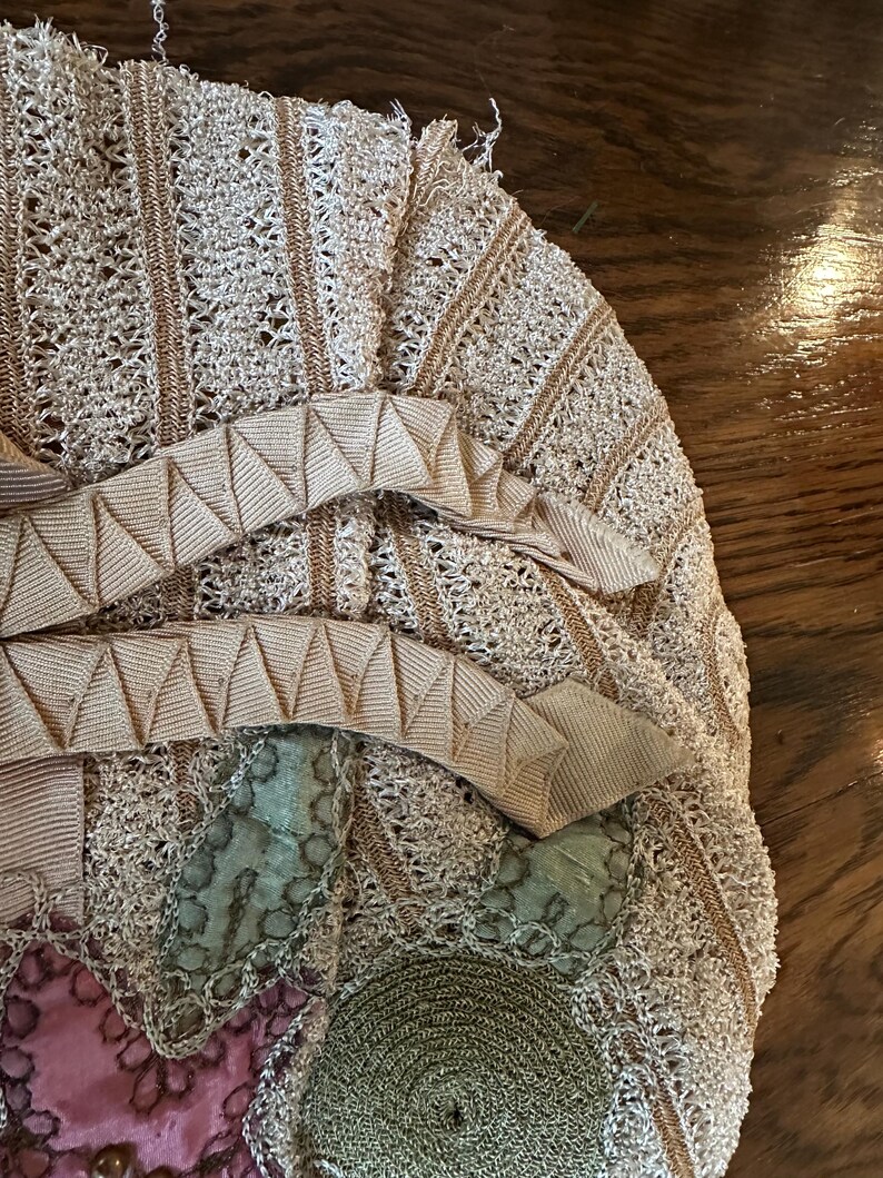 May include: Close-up of a vintage beige crocheted handbag with a decorative ribbon and floral accents. The bag features a detailed openwork design with a light brown trim and a pink and green floral pattern.