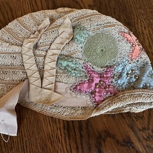 May include: An antique, beige-colored hat with a decorative design. The hat features a lace-like pattern, a ribbon, and colorful patches in pink, blue, and green. Small gold-colored beads are sewn onto the pink patch.