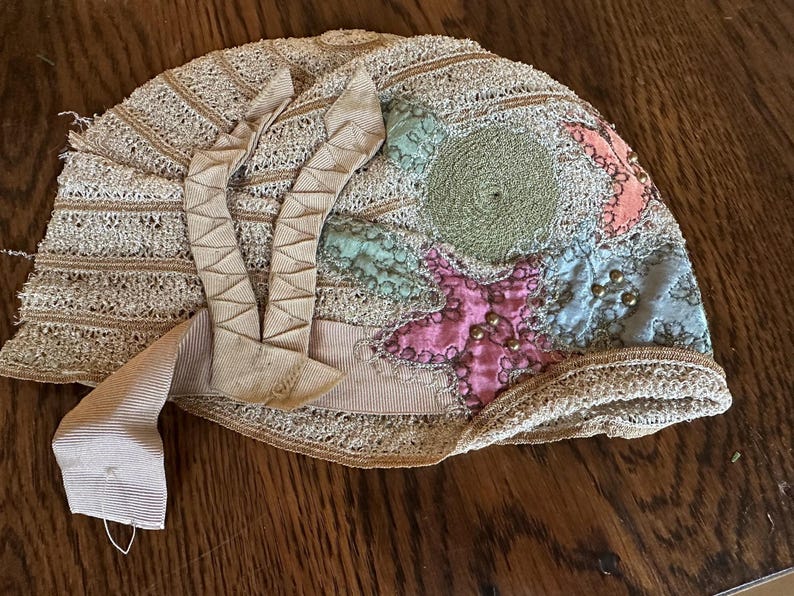 May include: An antique, beige-colored hat with a decorative design. The hat features a lace-like pattern, a ribbon, and colorful patches in pink, blue, and green. Small gold-colored beads are sewn onto the pink patch.