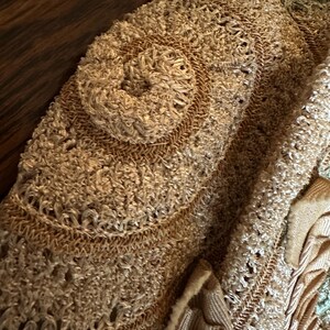 May include: Close-up of a beige, textured textile with a circular, raised design. The fabric appears to be hand-stitched or crocheted, with a mix of smooth and looped textures. Additional decorative elements are visible.