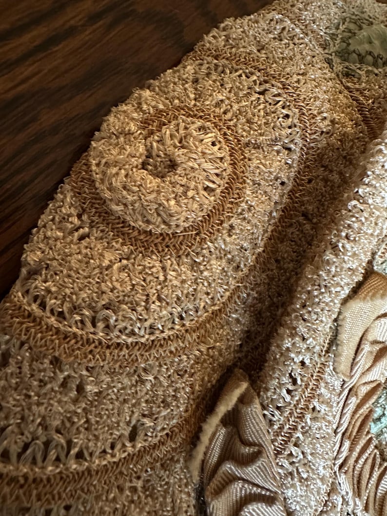 May include: Close-up of a beige, textured textile with a circular, raised design. The fabric appears to be hand-stitched or crocheted, with a mix of smooth and looped textures. Additional decorative elements are visible.