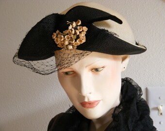 May include: A white and black straw hat with a black veil and a cluster of small, beige flowers on the side.