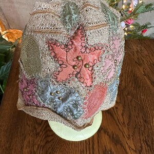 May include: A vintage-style hat with a patchwork design. The hat features a large pink star in the center, surrounded by patches of pink, blue, and green fabric. Gold-colored beads and trim add detail to the hat. The hat is displayed on a light green stand.
