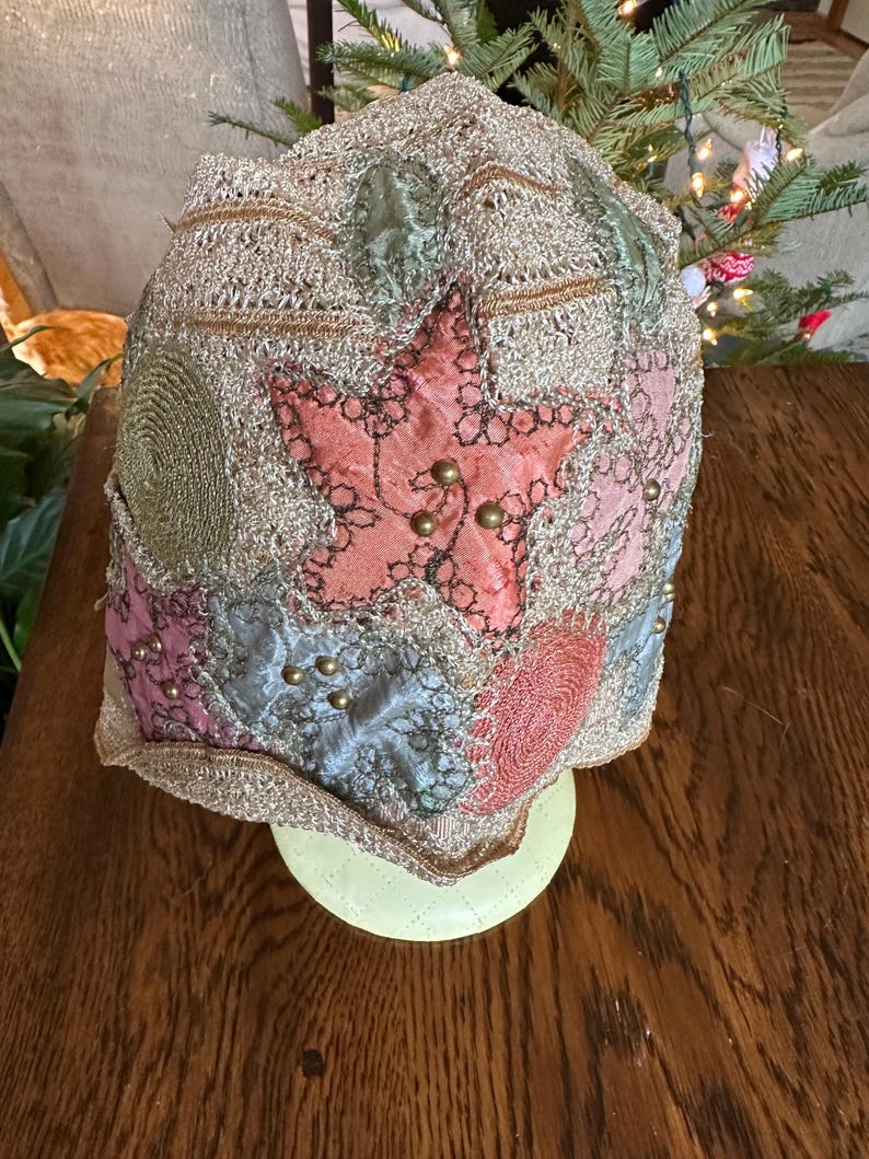 May include: A vintage-style hat with a patchwork design. The hat features a large pink star in the center, surrounded by patches of pink, blue, and green fabric. Gold-colored beads and trim add detail to the hat. The hat is displayed on a light green stand.