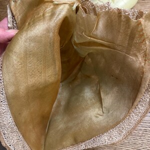 May include: A vintage, open-topped hat with a gold-colored silk lining and a delicate, cream-colored lace trim. The hat's interior is visible, showcasing the soft, lustrous fabric. The brim is edged with a fine, textured lace.