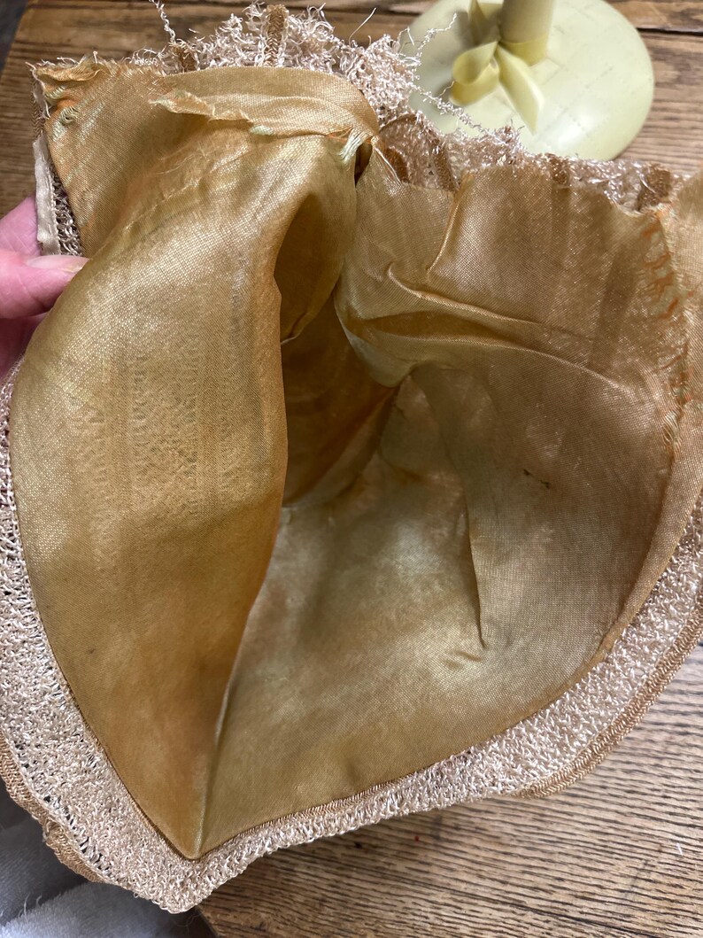 May include: A vintage, open-topped hat with a gold-colored silk lining and a delicate, cream-colored lace trim. The hat's interior is visible, showcasing the soft, lustrous fabric. The brim is edged with a fine, textured lace.