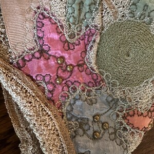 May include: Close-up of a vintage textile with a patchwork design. The fabric features pink, blue, and green velvet patches, embellished with gold-colored beads and intricate lace trim. A cream-colored ribbon is visible at the top.