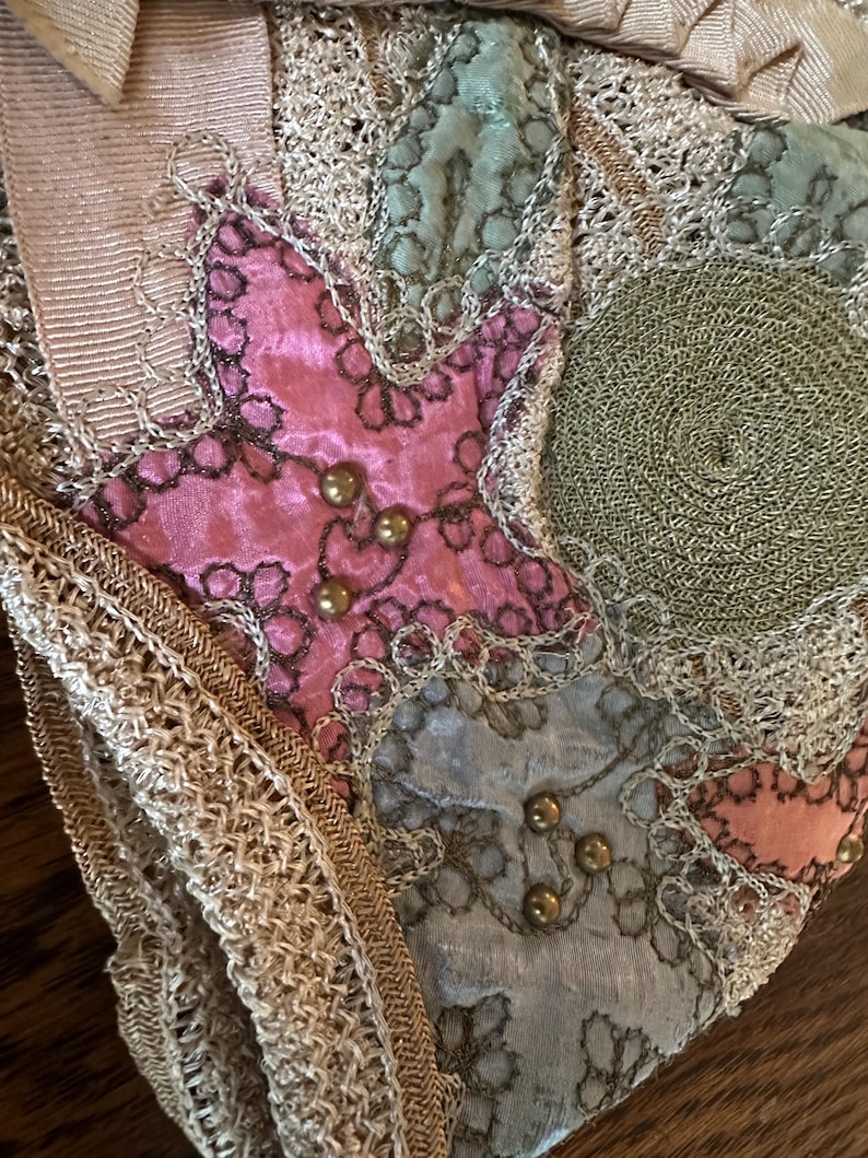May include: Close-up of a vintage textile with a patchwork design. The fabric features pink, blue, and green velvet patches, embellished with gold-colored beads and intricate lace trim. A cream-colored ribbon is visible at the top.