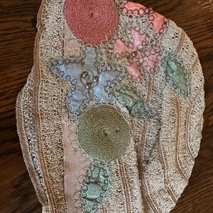 May include: Antique lace textile with floral embellishments. The piece features a delicate, openwork design in a light beige color. Appliqued with a pink, blue, and green floral design, and a pink ribbon. The textile is likely from the early 20th century.