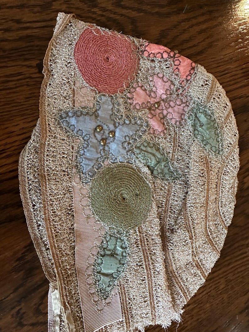 May include: Antique lace textile with floral embellishments. The piece features a delicate, openwork design in a light beige color. Appliqued with a pink, blue, and green floral design, and a pink ribbon. The textile is likely from the early 20th century.