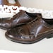 May include: A pair of brown leather lace-up shoes with a brogue design. The shoes have a black rubber sole and are worn.