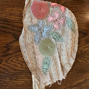 May include: An antique, off-white lace applique with colorful floral embroidery. The design features pink, blue, and green circular and leaf-shaped elements, accented with gold beads and a pink ribbon.