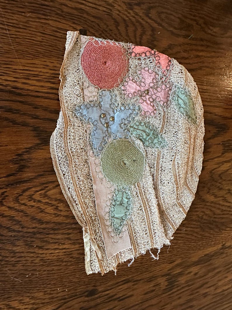 May include: An antique, off-white lace applique with colorful floral embroidery. The design features pink, blue, and green circular and leaf-shaped elements, accented with gold beads and a pink ribbon.
