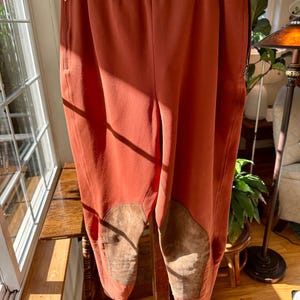 May include: Rust-colored riding pants with suede knee patches. The pants have a high waist, belt loops, and a zippered pocket on the side. The pants are hanging on a hanger.