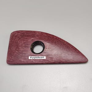 May include: A wooden pottery rib tool with a dark purple hue and a circular hole. The tool has a curved shape and the text "Purpleheart" is printed on a small white label.