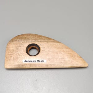 May include: A wooden pottery rib tool made of Ambrosia Maple. The tool is light brown with a dark brown circular hole. The text "Ambrosia Maple" is printed on a white label on the tool.