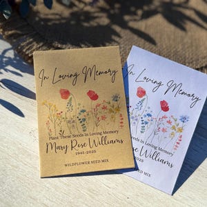 May include: Two seed packets with floral designs. One is tan, the other white. Both read "In Loving Memory" and "Mary Rose Williams 1945-2025". The tan packet says "Wildflower Seed Mix". The packets feature watercolor illustrations of red poppies and other wildflowers.