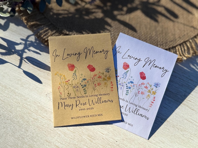 May include: Two seed packets with floral designs. One is tan, the other white. Both read "In Loving Memory" and "Mary Rose Williams 1945-2025". The tan packet says "Wildflower Seed Mix". The packets feature watercolor illustrations of red poppies and other wildflowers.